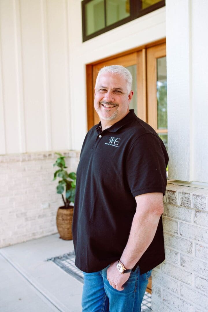 Smiling man in black polo shirt standing outside near a white wall.