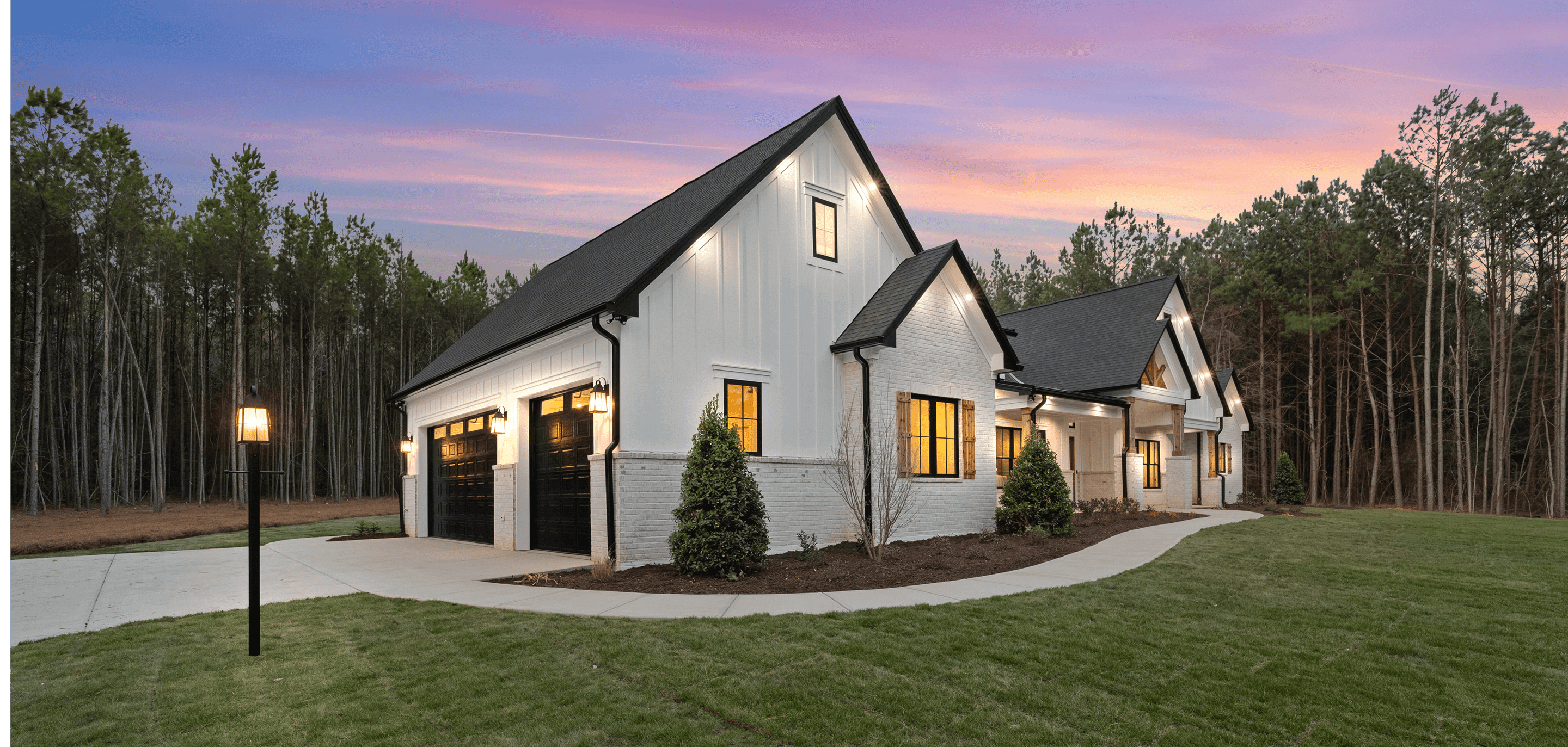 Modern white house with black roof and warm interior lighting at sunset.