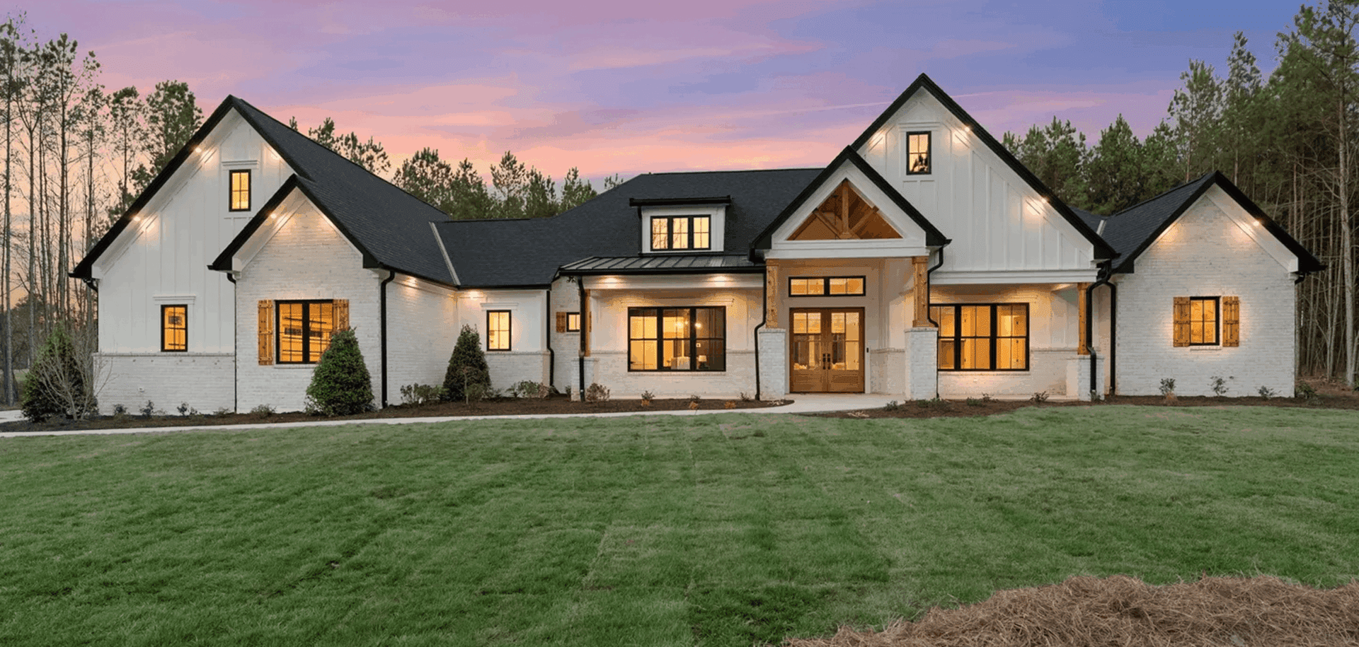 Modern farmhouse with warm lighting and a spacious lawn at dusk.